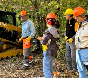 Workers in a forested area wearing hard hats and other safety equipment with a large piece of construction equipment in the background.